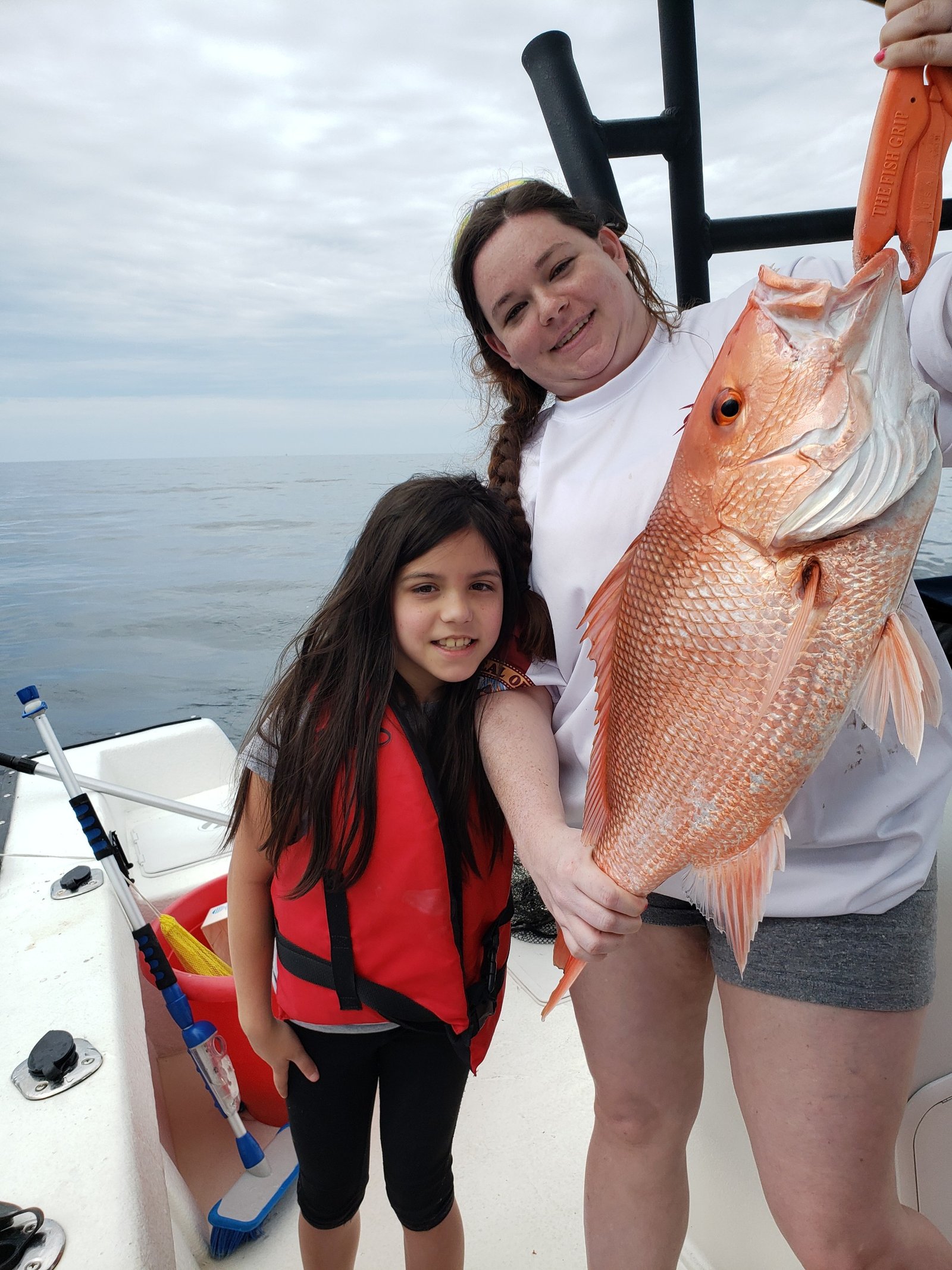An adult and a child on a boat; the adult is holding a large fish with an orange clamp while the child stands nearby wearing a red life jacket.